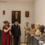Dwie osoby w strojach z epoki stoją w muzeum. W tle popiersia i portret na jasnych ścianach. Oświetlenie punktowe.