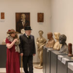 Dwójka ludzi w eleganckich strojach ogląda rzeźby w muzeum. Ściany są jasne z wiszącymi portretami. Tło jest minimalistyczne.