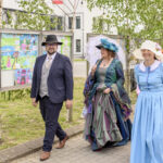 Osoby w strojach historycznych idą chodnikiem, na tle zielonych drzew. Mężczyzna w garniturze i kapeluszu. Kobiety w barwnych sukniach. Tło z kolorowymi rysunkami na tablicy.