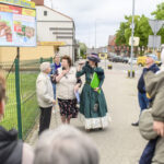 Grupa ludzi na chodniku, w tle billboard reklamowy. Kobieta w kostiumie z epoki trzyma mikrofon. Obok zielony płot i drzewa.