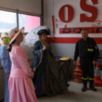 Grupa osób w strojach retro i strażak. Ściana z logo OSP w tle. Jasne światło wpada przez duże okno.