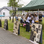 Na zdjęciu jest zielony namiot z ludźmi w białych koszulkach. Obok stoi wystawa z dużymi czarno-białymi plakatami. W tle widać biały budynek i niebieski samochód.
