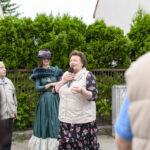 Trzy osoby w rozmowie na tle zielonych krzewów. Kobieta trzyma mikrofon, ubrana w kwiecistą sukienkę. Jedna z osób ma strój w stylu retro.