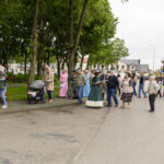 Na zdjęciu grupa ludzi spaceruje po ulicy. Część z nich jest ubrana w historyczne stroje. W tle widać drzewa i budynki.