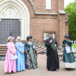 Grupa osób w kolorowych strojach historycznych stoi przed ceglaną fasadą kościoła. Na środku mężczyzna w czarnym stroju, trzyma mikrofon. Tło stanowi drzwi kościoła i zieleń po prawej stronie.