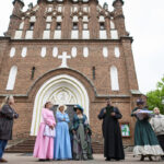 Kościół z cegły, ludzie w strojach historycznych przed wejściem. Jasne kolory sukien. Tło zielone drzewa.