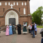 Grupa ludzi w strojach historycznych stoi przed ceglanym kościołem z białymi detalami. Przewodnik w czarnej szacie przemawia. W tle zielone drzewa.