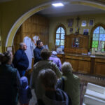 Grupa ludzi w pomieszczeniu kościelnym z drewnianymi meblami i kolorowymi witrażami. Na ścianie wiszą obrazy i krzyż. W tle widać figury religijne.