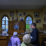 Wnętrze pokoju z drewnianymi szafkami, ozdobione zdjęciami i krzyżem. Ludzie oglądają ścianę z kolorowymi witrażami. Atmosfera religijna, spokojna.