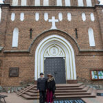 Para stoi przed zabytkowym, ceglastym kościołem. Widoczne schody i duże, zdobione drzwi. Strój elegancki, tło z czerwonej cegły.