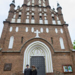 Ceglana fasada kościoła z licznymi ozdobnymi oknami. Przed wejściem stoi elegancka para. Na ziemi napis PARAFIA.