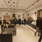 Wnętrze muzeum z rzeźbami na ciemnych podestach. Kilka osób ogląda eksponaty w jasnym pomieszczeniu. Sufit ozdobiony reflektorami.