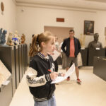 Dziewczynka w czarnej kurtce pisze na kartce w galerii sztuki. Wokół niej rzeźby i kilka osób. Jasne, eleganckie wnętrze.