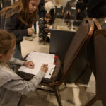 Dwie dziewczynki rysują na kartkach przy ciemnych krzesłach. W tle widać ludzi w muzeum. Dominuje ciepła, brązowa kolorystyka.