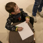 Chłopiec w kurtce z logo nietoperza rysuje na białej kartce. Siedzi na brązowym krześle. Tło to jasne płytki podłogowe.