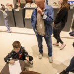 Chłopiec rysuje na kartce w galerii sztuki. Dorośli wokół oglądają eksponaty na szarych postumentach. Podłoga jest jasnobrązowa.