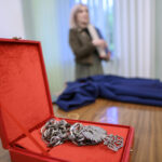 Czerwone pudełko z metalowym łańcuchem leży na drewnianym stole. W tle nieostro widoczna osoba i niebieska tkanina. Jasne ściany dopełniają scenerię.