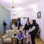 Pokój z jasnymi ścianami, trzema żyrandolami, zielonymi zasłonami. Ludzie siedzą i stoją wokół foteli i sofy. Na ścianie obraz.