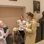 Cztery osoby rozmawiają w muzeum, otoczone rzeźbami i obrazami. Ściany są jasne, a osoby trzymają kartki. Pomieszczenie jest dobrze oświetlone.