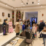 Sala muzealna z popiersiami na ścianach, kolorowe palmy w rogu. Na środku grupa osób, niektóre w historycznych strojach. Jasne oświetlenie, beżowe ściany.