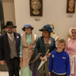 Grupa osób w strojach retro pozuje w muzeum z popiersiami w tle. Dominują kolory: beżowy, niebieski i różowy. Ściany są białe, eksponaty ustawione na ciemnych stojakach.