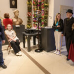 Grupa osób w muzeum, z rzeźbami i ścianą kwiatów w tle, jasne wnętrze. Mężczyzna w meloniku i kobieta z futrem.