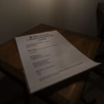 Arkusz papieru na drewnianym stole z tekstem w kolorze niebieskim. Oświetlenie jest przyciemnione, co nadaje ciepły ton. Zdjęcie skupia się na treści dokumentu.