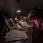 Osoba w różowej sukni pisze przy biurku, oświetlona lampką. Na stole leżą dokumenty i zdjęcia w ramkach. Tło jest ciemne.