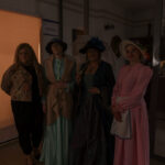 Cztery kobiety w strojach vintage stoją w pomieszczeniu. Jedna trzyma parasolkę, a inna ma różową suknię. Tło ciemne, osłonięte roletą.