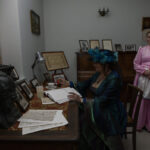 Na zdjęciu dwie kobiety w historycznych strojach siedzą i stoją w eleganckim pomieszczeniu z biurkiem. Kobieta siedząca pisze w księdze, ubrana w ciemnozieloną suknię z piórami na kapeluszu. Tło zdobi