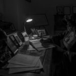 Ciemny pokój z biurkiem, na którym kobieta w kapeluszu pisze w notatniku. Lampka oświetla zdjęcia na stole. Obok stoi maszyna do pisania.