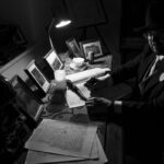 Czarno-białe zdjęcie biurka z mężczyzną piszącym przy lampce. Obok są ramki ze zdjęciami i maszyna do pisania. Scena przypomina dawny gabinet.