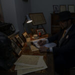 Mężczyzna w kapeluszu pisze przy drewnianym biurku, otoczony zdjęciami i maszyną do pisania. Światło lampy tworzy ciepły klimat. Tło stanowi ciemne wnętrze z drewnianym meblem.