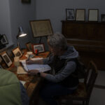 Osoba przy biurku pisząca w notatniku, otoczona starymi zdjęciami. Światło lampki biurkowej oświetla przestrzeń. W tle brązowe pianino z ramkami.