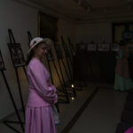 Kobieta w różowej sukni i kapeluszu stoi w galerii, oglądając czarno-białe zdjęcia na sztalugach. Tło jest ciemne. Obok siedzi osoba w różowym szalu.