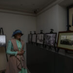 Pomieszczenie z czarno-białymi zdjęciami na stojakach. Kobieta w stroju retro, w kapeluszu, stoi z lewej strony. Jasne, neutralne tło.