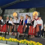 EcZqNGqA Grupa ludzi w strojach ludowych śpiewa na scenie. Mają czerwone i kolorowe spódnice oraz kamizelki. Tło zdobi niebieskie zadaszenie, a przód żółte kwiaty.