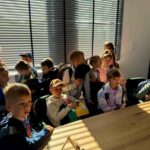 Grupa dzieci z plecakami siedzi w jasnym pokoju, ciepłe światło pada przez żaluzje. Pomieszczenie ma drewniany stół. Dzieci patrzą w stronę aparatu.