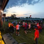 Kj02zJmw Impreza plenerowa w parku, ludzie tańczą na trawie. Wieczorne niebo z chmurami, scena oświetlona, żółte kwiaty na scenie. W tle widoczne namioty i publiczność.