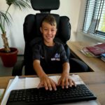 Chłopiec siedzi przy biurku z klawiaturą, uśmiechnięty na czarnym fotelu. W tle jest jasna ściana i roślina w doniczce. Obok biurka leżą segregatory i dokumenty.