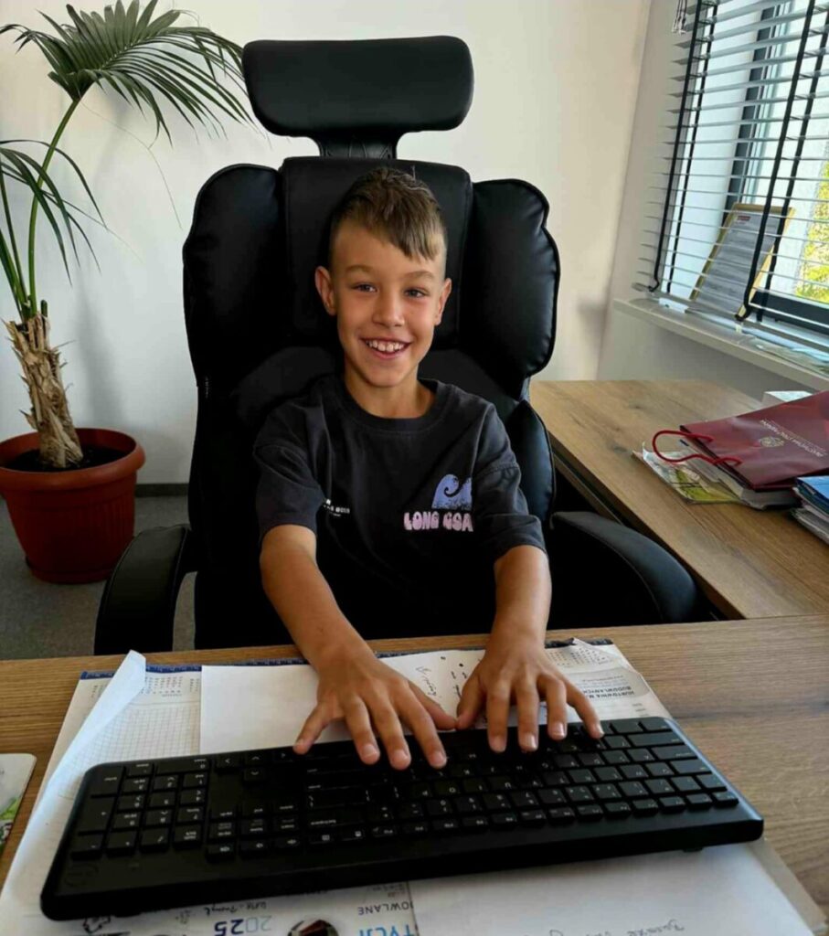 Chłopiec siedzi przy biurku z klawiaturą, uśmiechnięty na czarnym fotelu. W tle jest jasna ściana i roślina w doniczce. Obok biurka leżą segregatory i dokumenty.