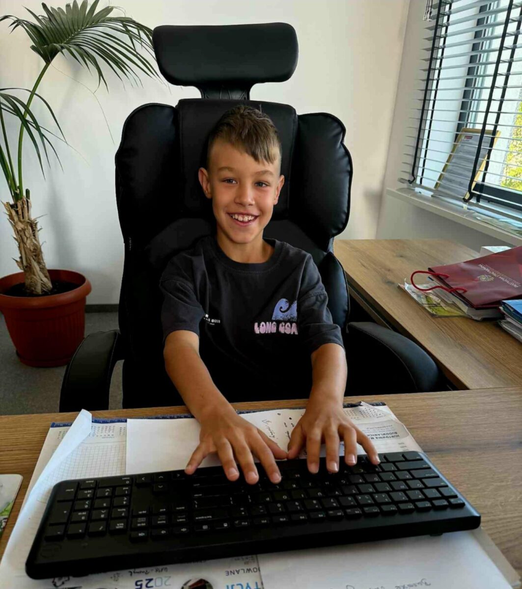 Chłopiec siedzi przy biurku z klawiaturą, uśmiechnięty na czarnym fotelu. W tle jest jasna ściana i roślina w doniczce. Obok biurka leżą segregatory i dokumenty.