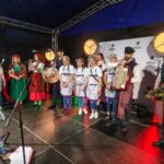 QRyjSa5T Ludzie w kolorowych, tradycyjnych strojach ludowych stoją na scenie. Jasne światła reflektorów oświetlają grupę. W tle ciemne niebo i białe tło.