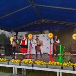 UGDHQUMc Grupa muzyków w tradycyjnych strojach na scenie pod niebieskim zadaszeniem. Tło z czarnym ogrodzeniem, scenę zdobią żółte kwiaty. Jasne reflektory oświetlają zespół.