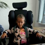 Dziewczynka siedzi na czarnym fotelu biurowym. Ma na sobie biało-różową bluzę z czarnymi wzorami. W tle jasna ściana i okno z żaluzjami.