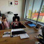 Dwoje dzieci w biurze; jedno siedzi na czarnym fotelu przy drewnianym biurku z klawiaturą. Biel ścian, godła, roślina i okno z żaluzjami. Na biurku zdjęcie w ramce i segregatory.