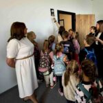 Grupa dzieci z plecakami stoi w pokoju z białymi ścianami, patrząc na kobietę dzwoniącą dzwonkiem. Obok dzieci stoją dwie kobiety. W tle znajdują się drewniane drzwi i roślina na górnej półce.