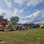 gqiuY60t Kolorowe dmuchane zjeżdżalnie stoją na zielonej trawie. Kilkanaście osób spaceruje wokół atrakcji. Na tle jasnego nieba namioty stoisk.