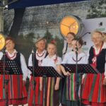 mLxaxhvO Grupa osób w ludowych strojach występuje na scenie z mikrofonami. Mają kolorowe spódnice i korale. Tło jest ciemne z reflektorami.