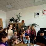 Grupa dzieci w biurze w jasnym pomieszczeniu. Na ścianie wisi godło Polski. W tle rośliny i drewniana szafka.